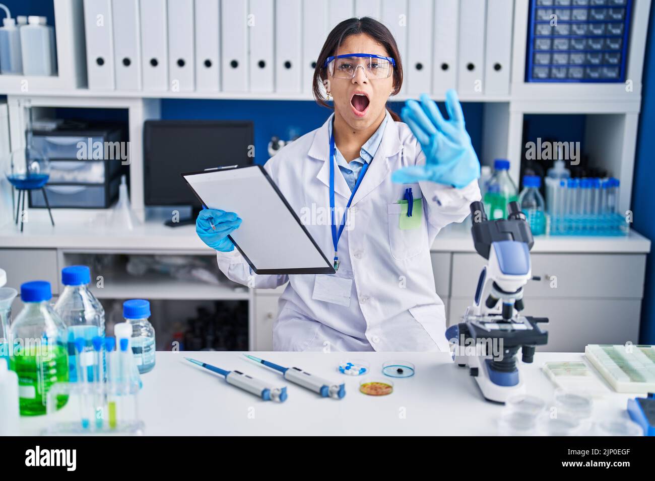 Hispanic young woman working at scientist laboratory doing stop gesture ...