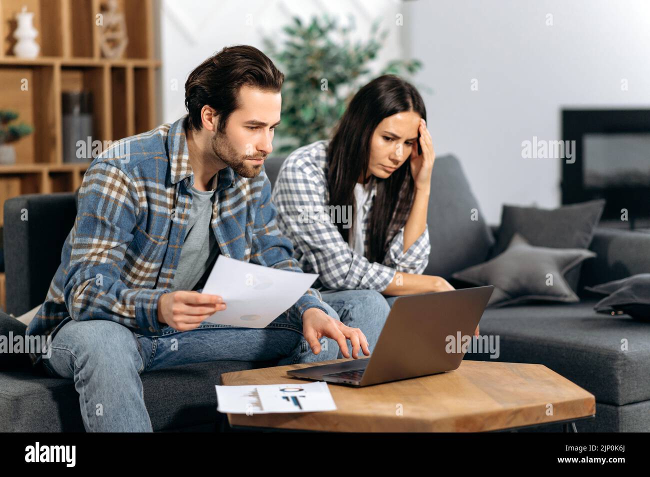 Frustrated caucasian couple sitting on the couch, the guy and the girl are freelancers, work from home, use a laptop, look at graphs, are unhappy with the results, low profit, thinking about strategy Stock Photo