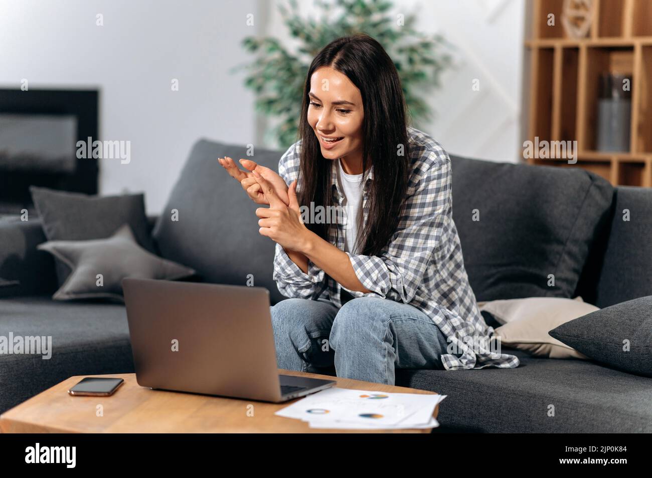 Positive caucasian successful brunette woman, freelancer or small business owner, sit on a sofa in the living room, using a laptop to work from home, communicate with colleague by video call, smiles Stock Photo