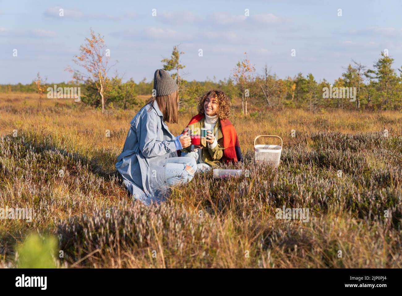 Smiling adult sisters make picnic on grass meadow after gathering berries against trees Stock Photo