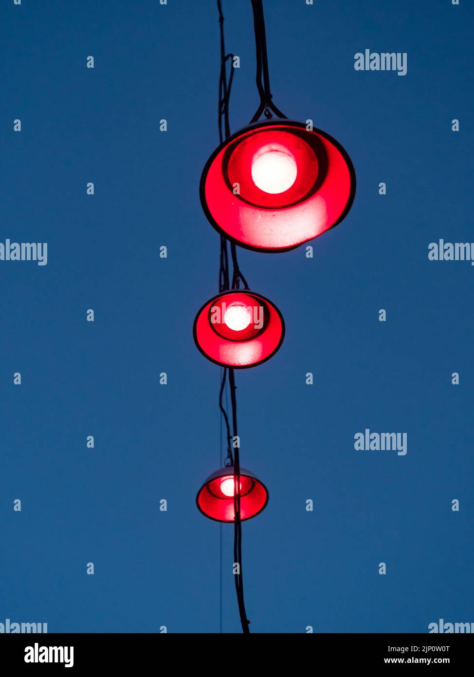 Three lamps hanging on cables in front of a blue evening sky. Red light is shining out of the light bulbs. An abstract scene with red street lights. Stock Photo