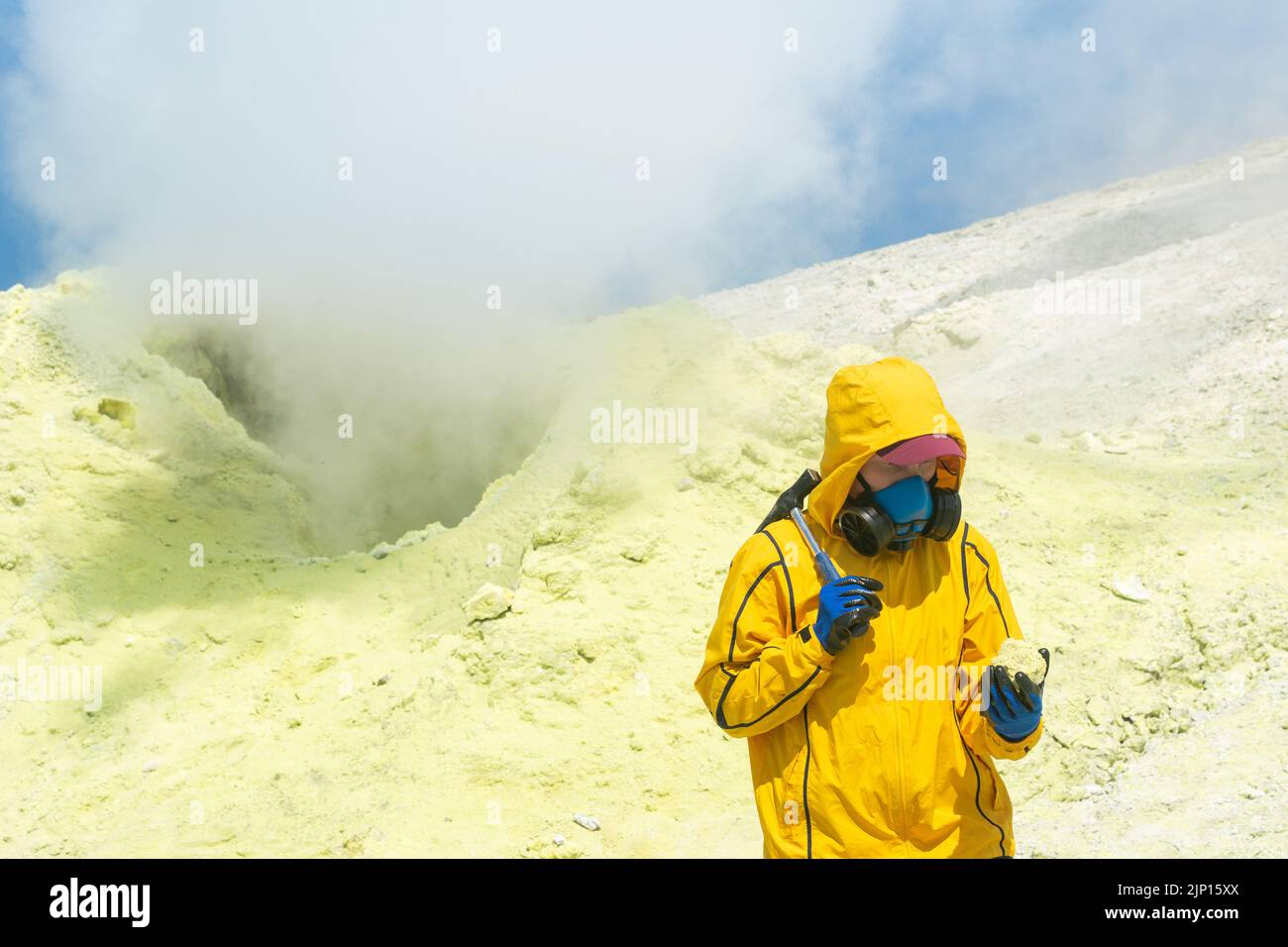 female volcano scientist on the background of a smoking fumarole examines a sample of a sulfur mineral Stock Photo