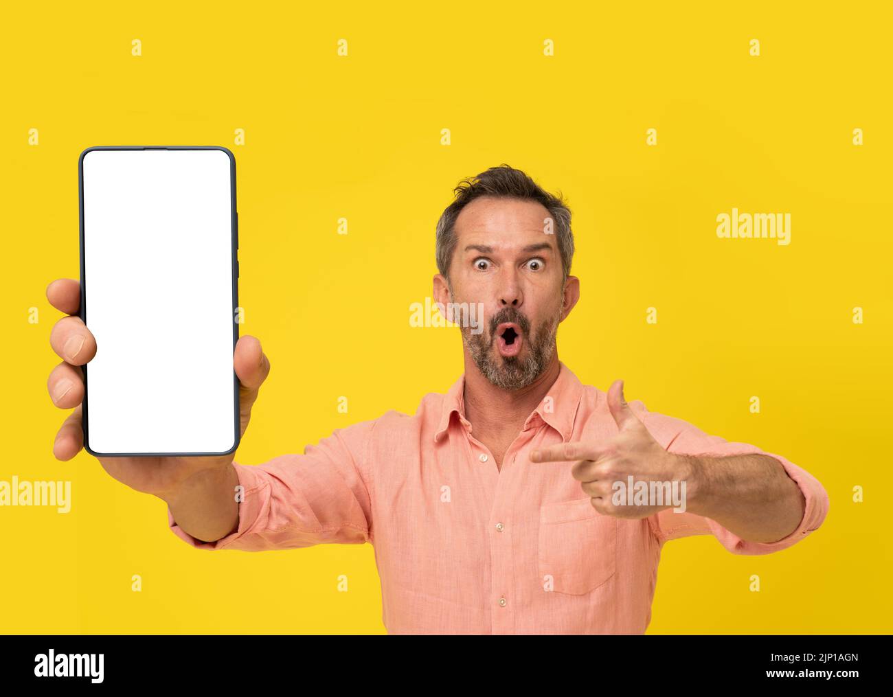 Amazed middle aged grey haired man pointing finger at smartphone looking at camera wearing peach shirt isolated on yellow. Mature fit man with phone app advertisement. Mock up white screen.  Stock Photo