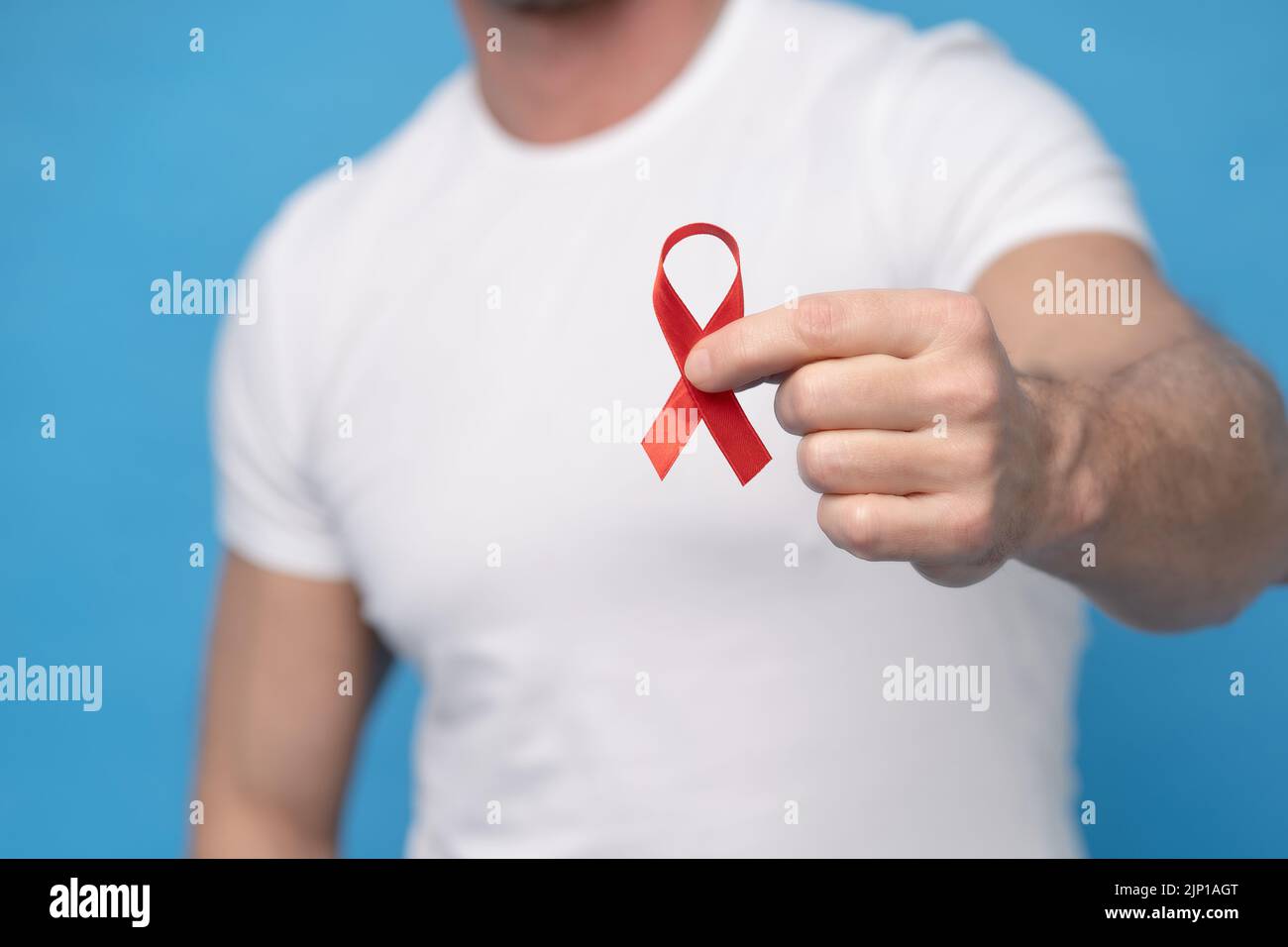 Man with red ribbon bow AIDS awareness symbol in hand wearing a white t-shirt isolated on a blue background. Modern medicine and healthcare. AIDS awareness concept. No face visible. Stock Photo