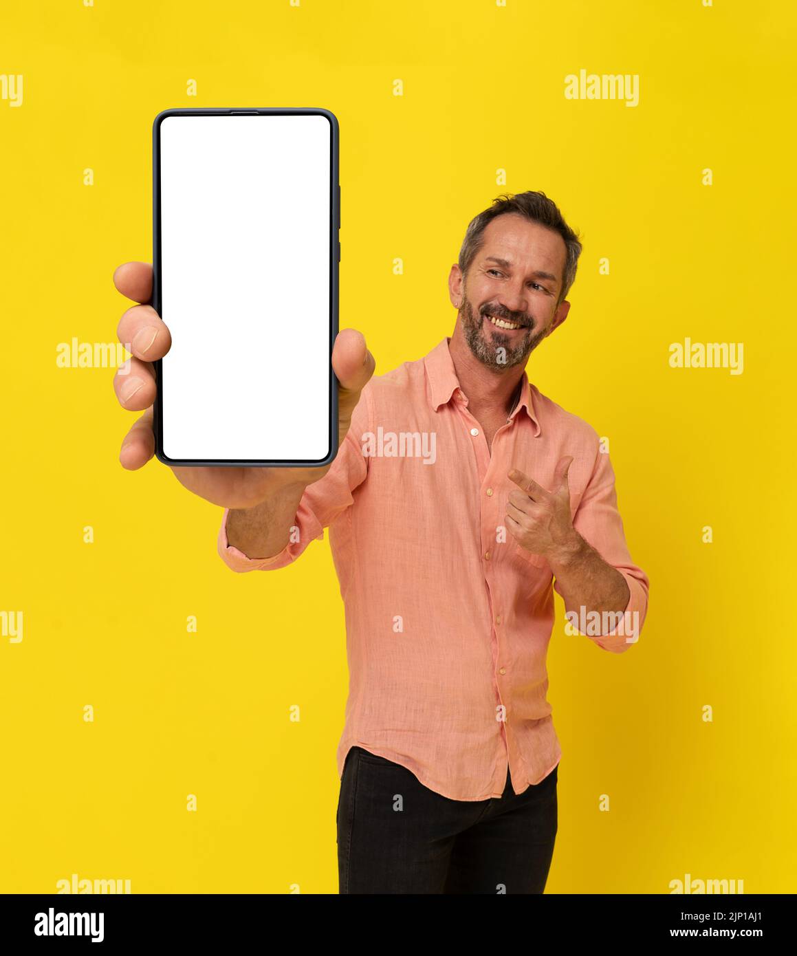 Happy middle aged grey haired fit man holding big smartphone pointing finger at it wearing peach shirt isolated on yellow. Mature muscled man with phone app advertisement. Mock up white screen.  Stock Photo
