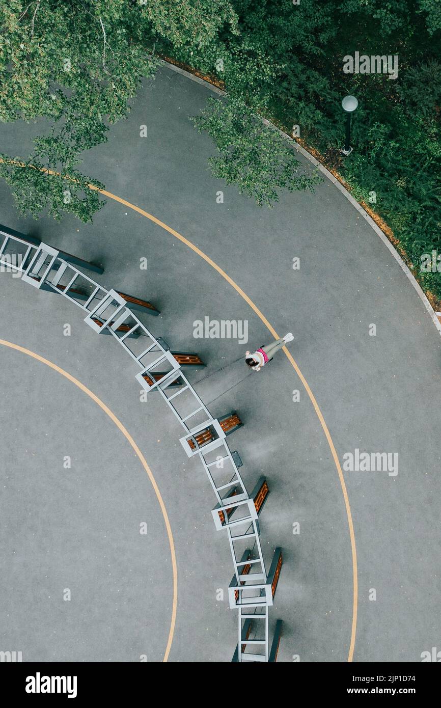 Lonely girl swings on hanging swing in park. Aerial drone view of seesaw in city park. Young caucasian woman in pants and tshirt swing up on curved swings in green summer park. Moody top down vertical Stock Photo