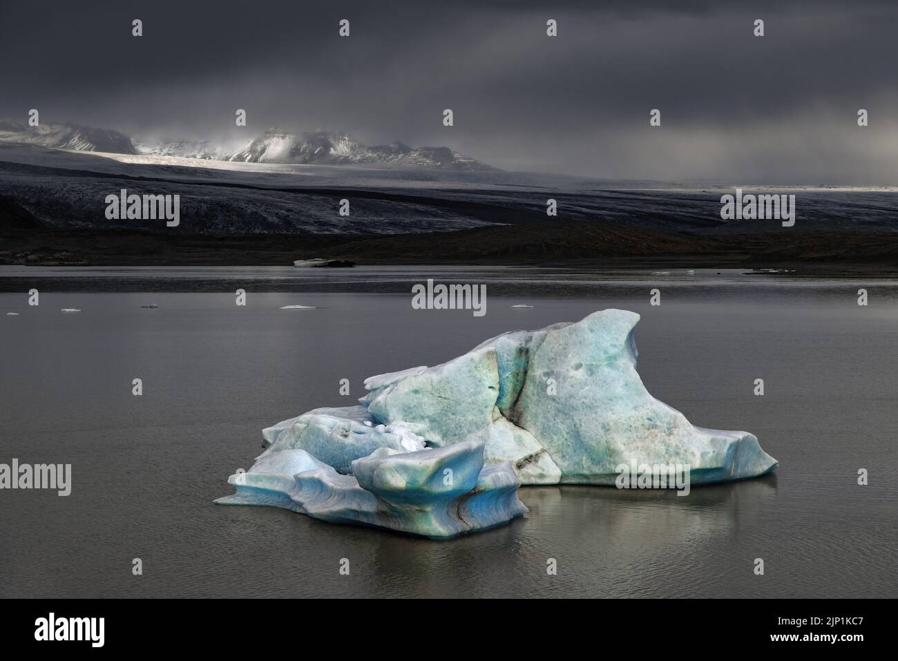 Fjallsarlon Glacier Lagoon, Iceland, on a stormy day. A blue iceberg floats in the lagoon and sunlight highlights the mountains behind. Part of the Va Stock Photo
