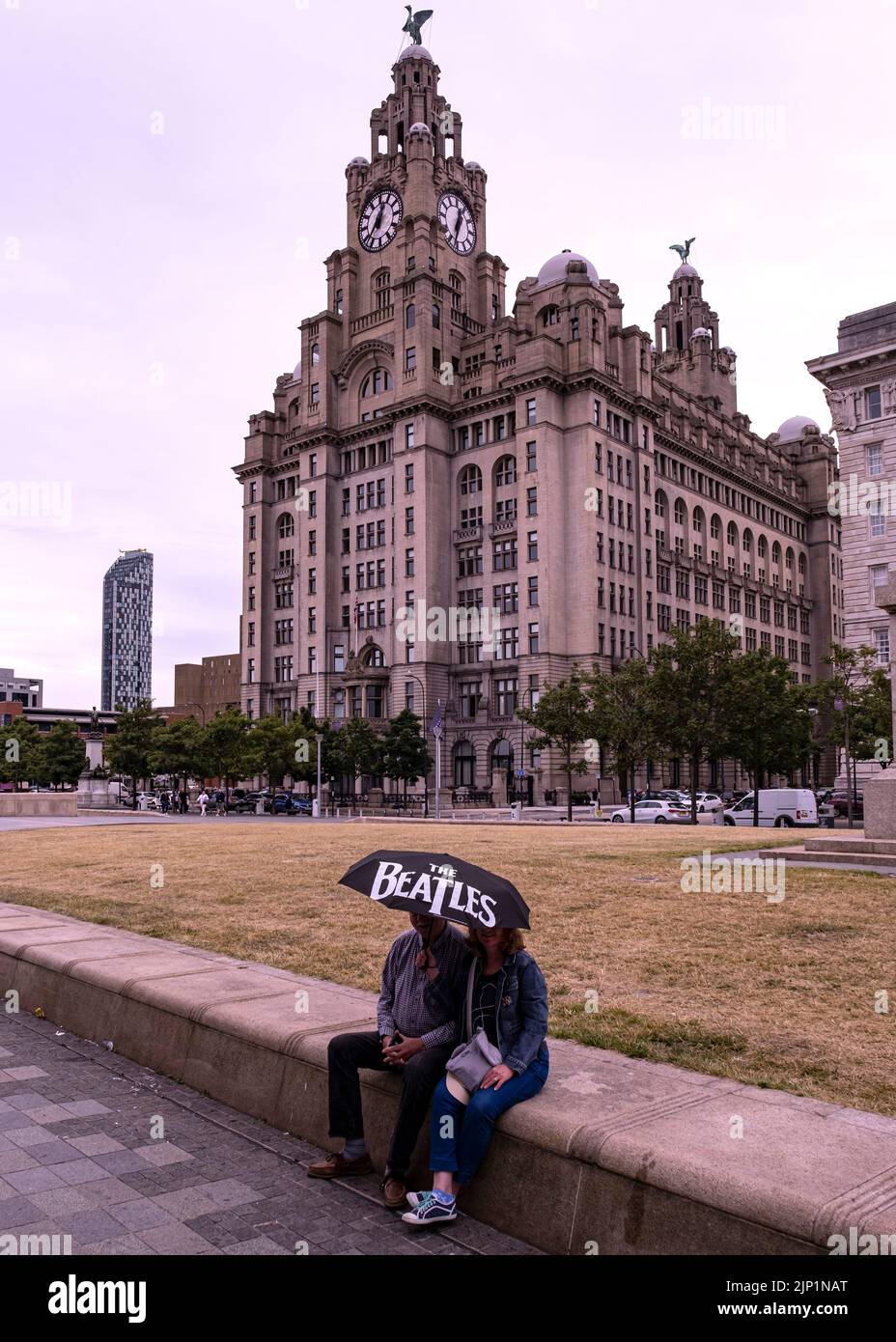 A couple sitting on a low wall with a Beatles umbrella, in front of the famous Royal Liver Building Stock Photo