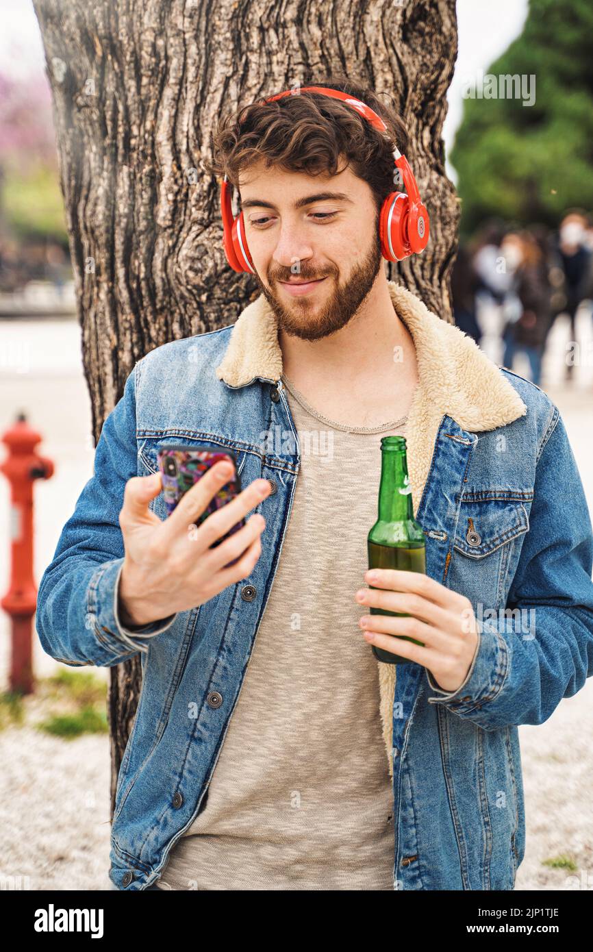 Young hipster guy listening to the music with wireless headphones outdoors or watching music media video with his smart mobile phone drinking a beer o Stock Photo
