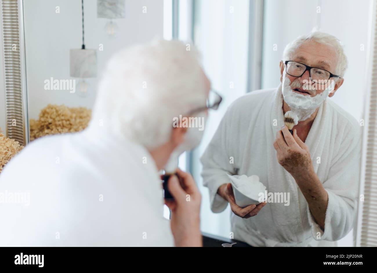 Senior man shaving his beard in the bathroom, looking in the mirror. Morning routine concept. Stock Photo