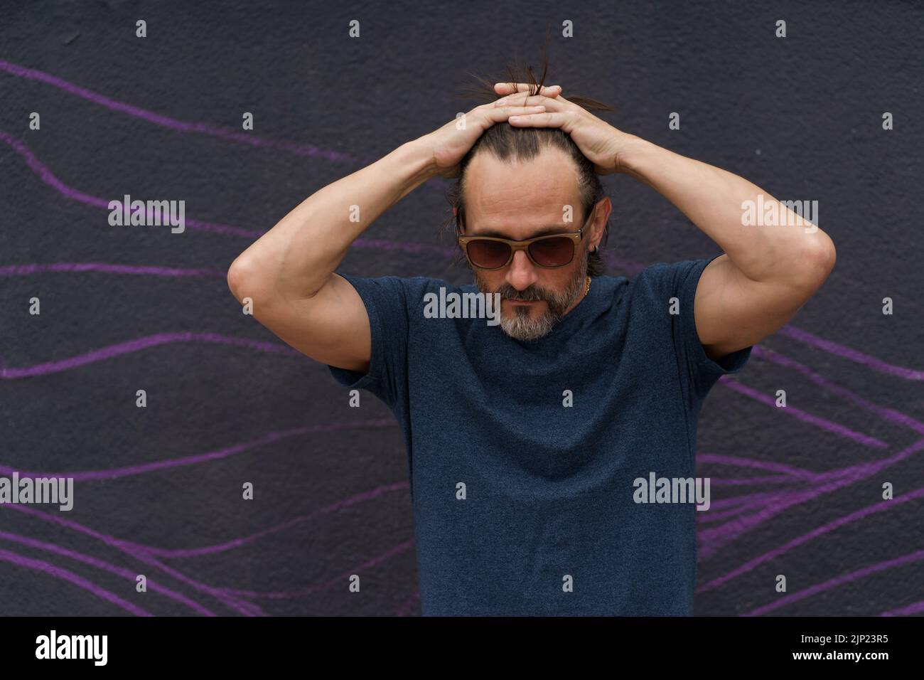 Thoughtful mature man standing with hand folded above his head next to dark urban wall leaned head down wearing sunglasses, dark blue t-shirt and light shorts. Stress, middle age crisis man concept.  Stock Photo