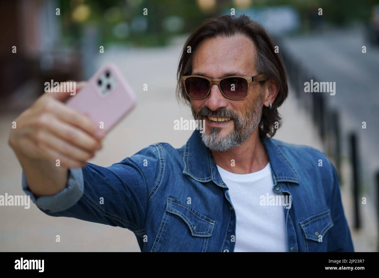 Man with smartphone outdoors making selfie or recording video message wearing denim shirt and sunglasses. Handsome middle aged man with grey hair making photos while traveling.  Stock Photo