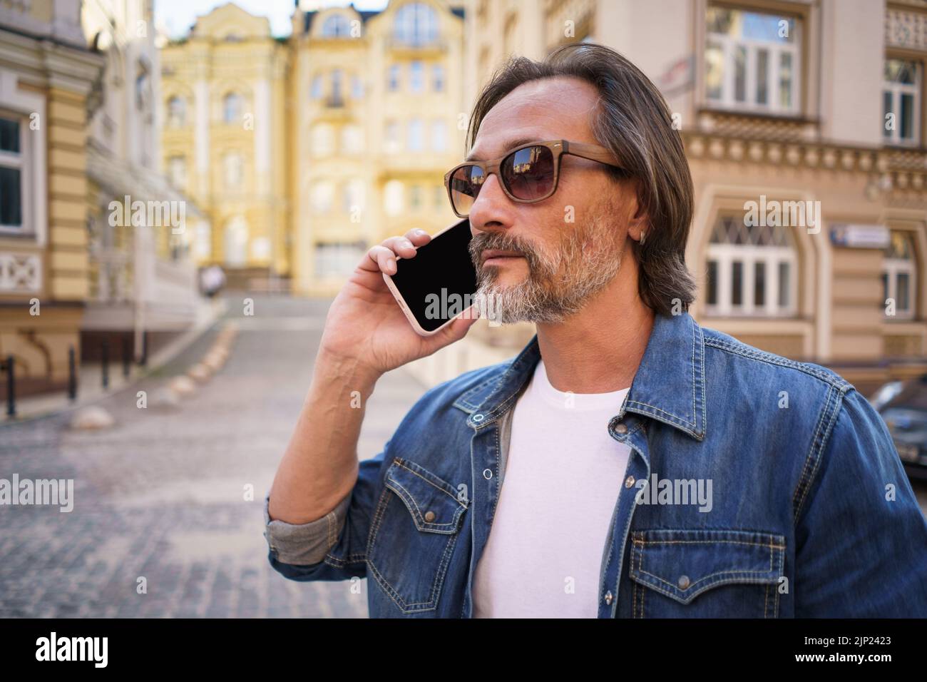 Middle aged man with grey bearded talking on the phone standing outdoors in old city background wearing denim jeans shirt. Mature business man working on the go. Freelancer traveling man.  Stock Photo