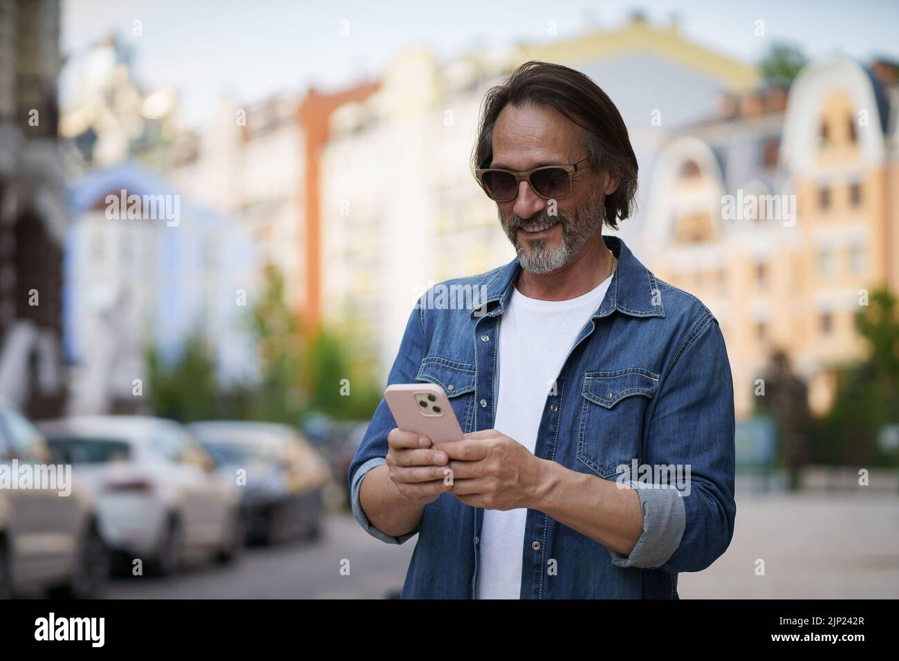 Texting or posting in social media post, photo mature handsome man with smartphone outdoors wearing denim shirt and sunglasses. Handsome middle aged man with grey hair making photos while traveling.  Stock Photo