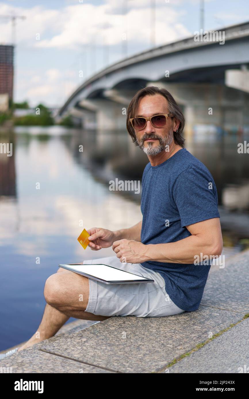 Working outdoors freelancer man sitting next to river using digital tablet pc with white screen, holding debit or credit card shopping online or paying online banking. E-banking concept.  Stock Photo