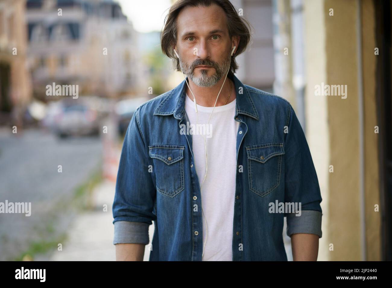 Handsome middle aged grey bearded man walking downtown with earphones listening music or talking on the phone wearing casual jeans shirt with white t-shirt. Travel lifestyle concept. Stock Photo
