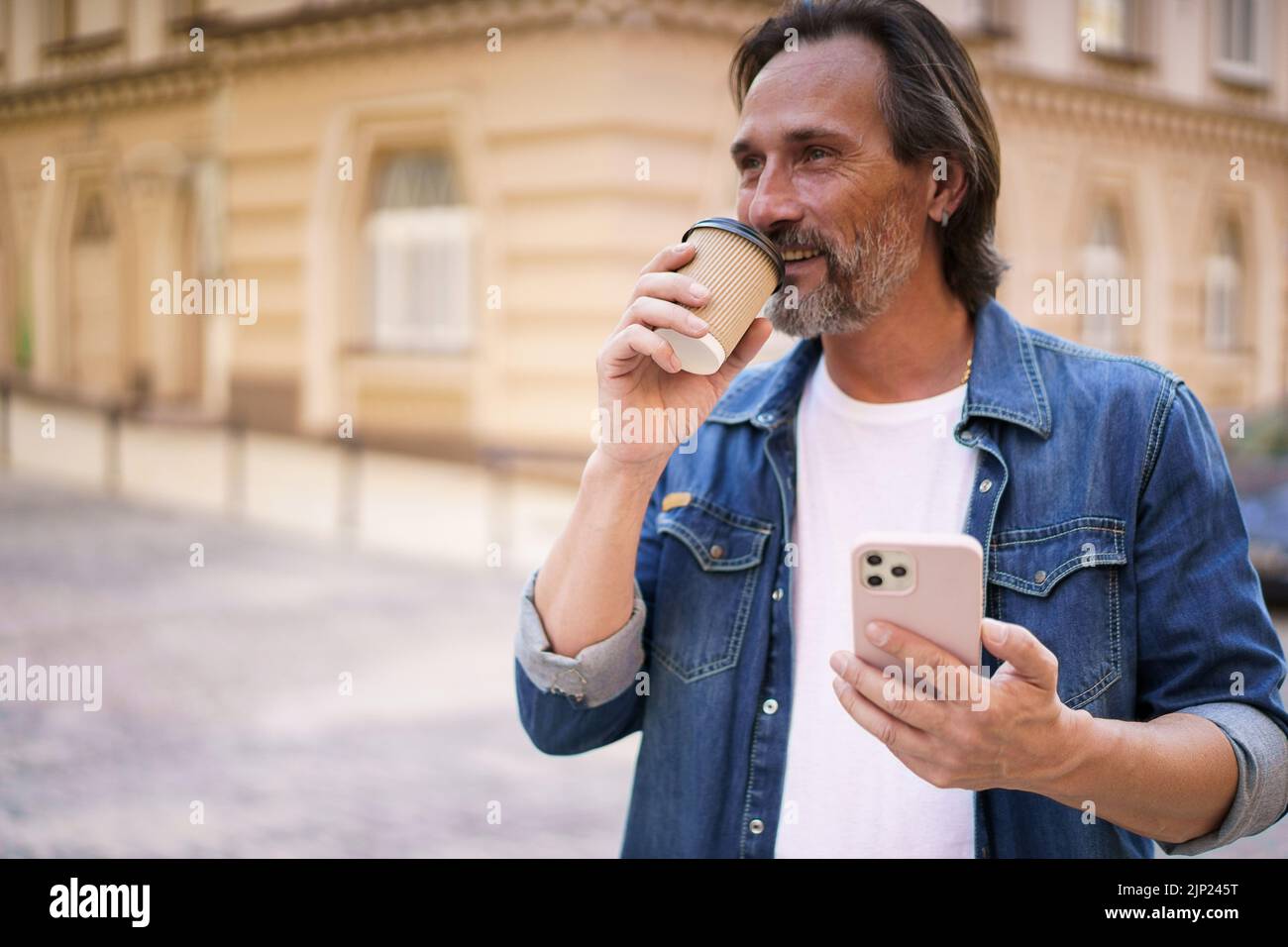 Mature man with coffee and smartphone outdoors. Middle aged man read having video call holding phone and take away paper cup with coffee wearing denim shirt. Business on the go concept.  Stock Photo