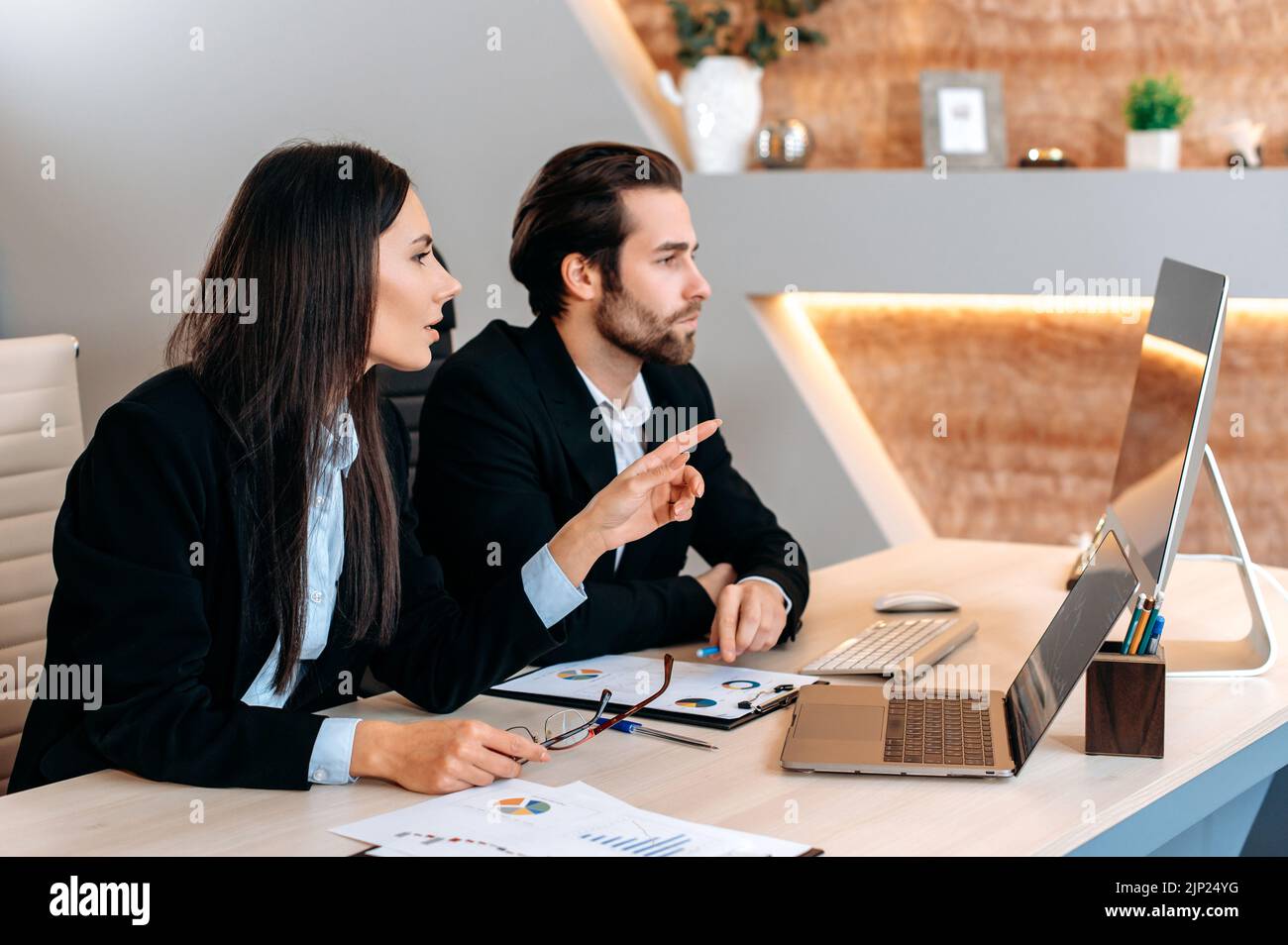 Two focused successful caucasian employees of the company, work together on a project while sitting at a table in a modern office, study financial charts, share information and ideas with each other Stock Photo