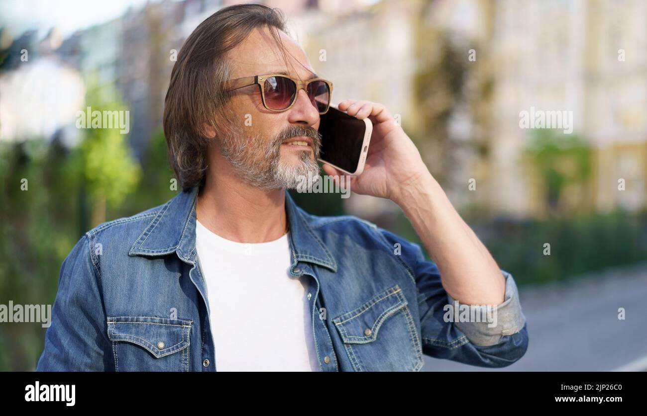 Mature bearded man talking on the phone standing outdoors in urban old city background wearing denim jeans shirt. Mature business man answering phone call standing outdoors.  Stock Photo
