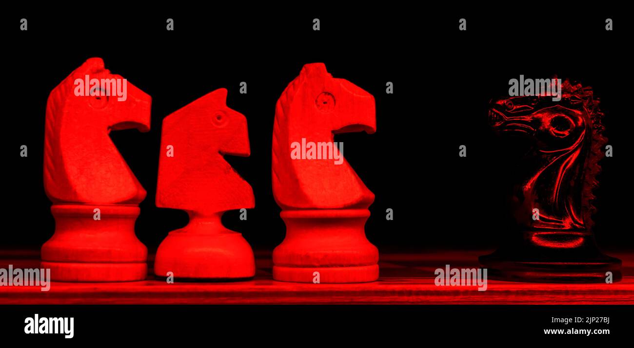 Black knight is standing on a chessboard against three white knights. Army of one. Selective focus. Neon red light in front of composition. Stock Photo