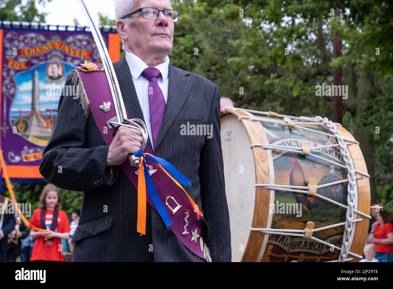 Antrim, 12th July 2022, UK. Man wearing crimson sash carrying a ceremonial sword walks ahead of lambeg drummers and Chaine's Inver Orange Lodge banner Stock Photo