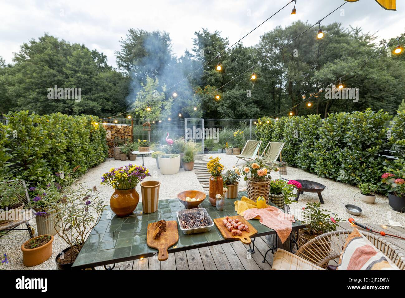 Atmospheric and cozy garden with dining place on terrace at dusk. Cooking food on disposable grill on beautiful table decorated with flowers Stock Photo
