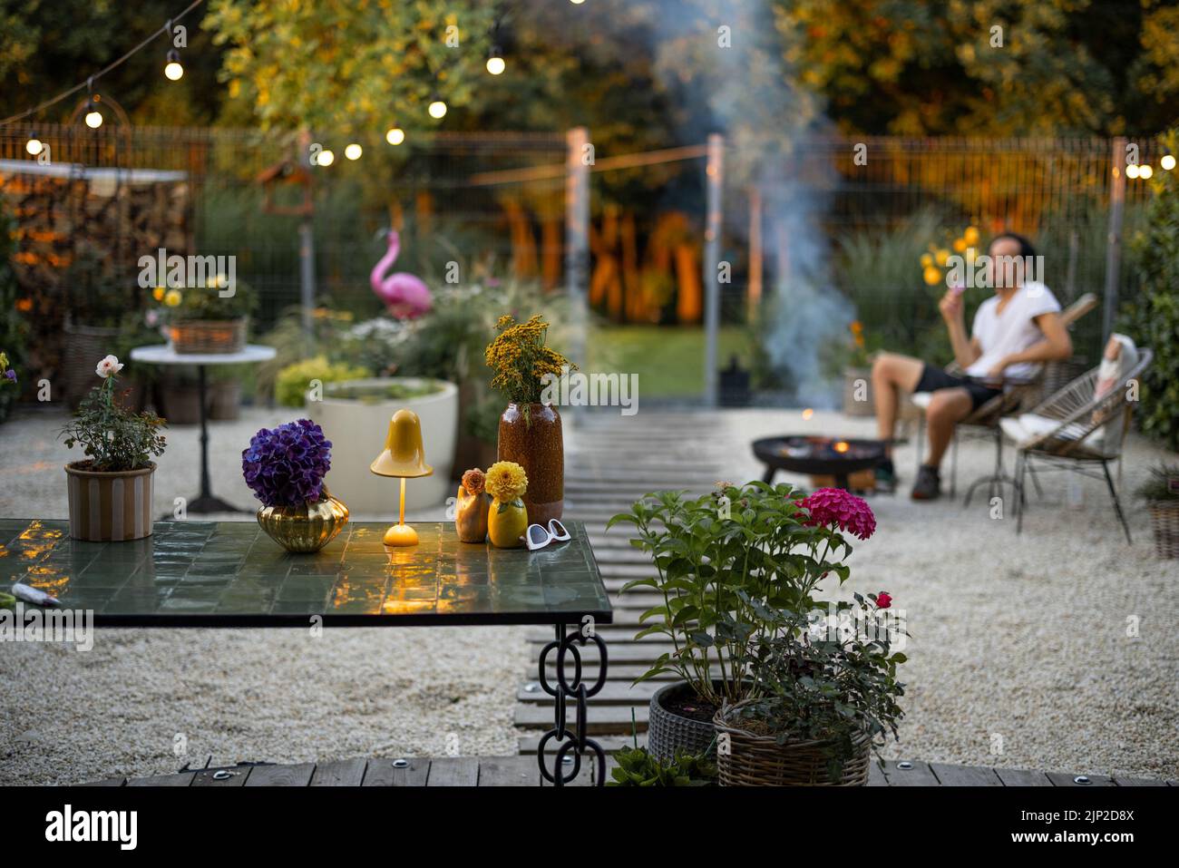 Beautiful and cozy garden with man relaxing sitting by the fireplace on background at dusk Stock Photo