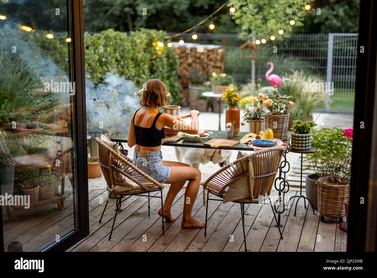 Woman cooks food on disposable grill while sitting relaxed by the table on cozy terrace during the evening at beautiful backyard Stock Photo