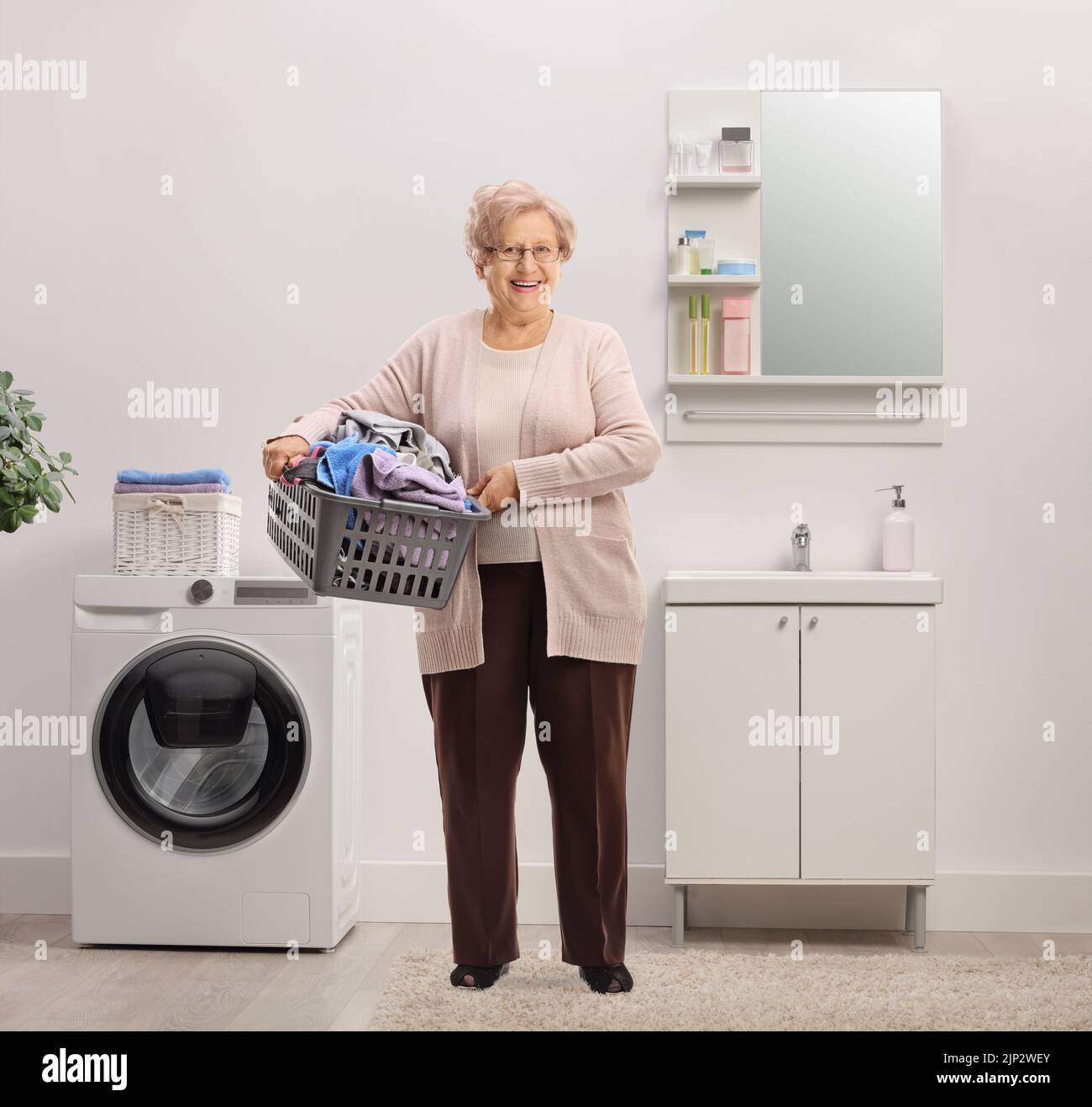 Full length portrait of an elderly woman holding a laundry basket with clothes inside a bathroom Stock Photo