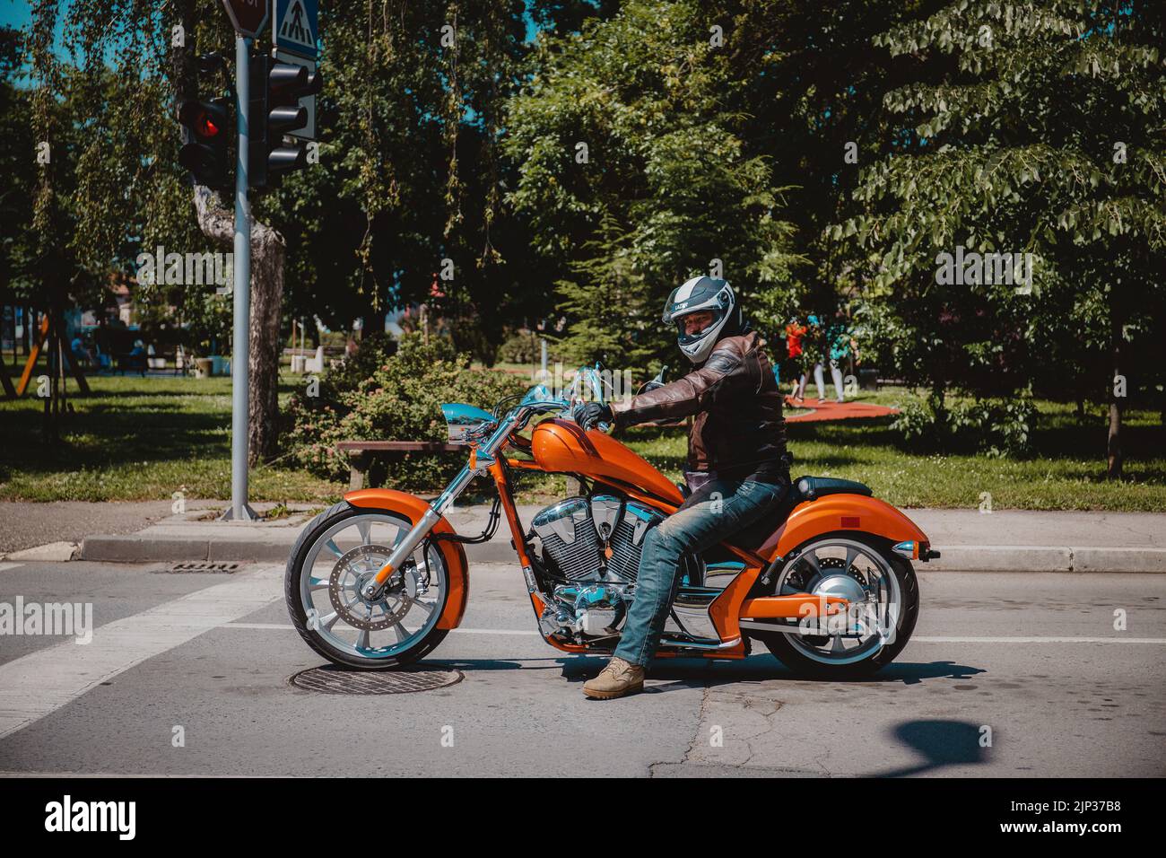 A biker with a helmet on a beautiful orange motorcycle on the road with green trees in the background Stock Photo