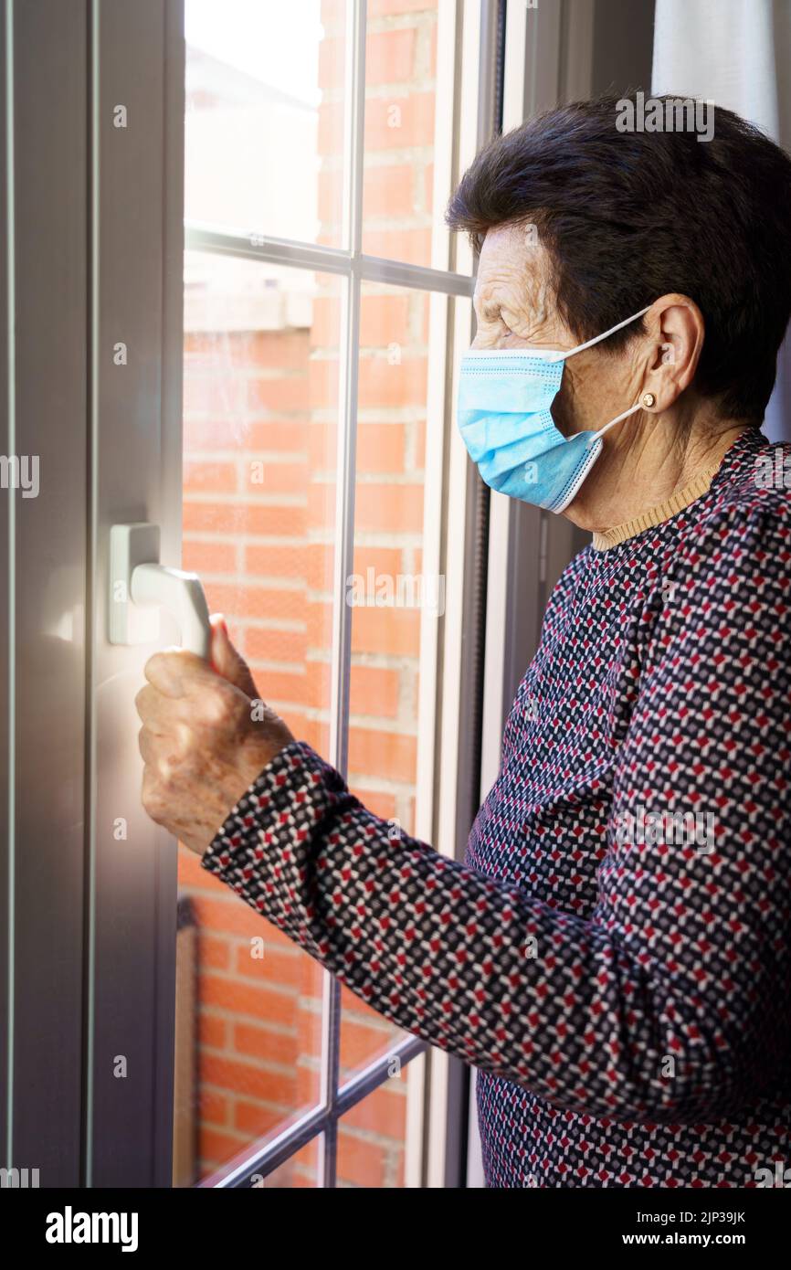 Elderly woman with a mask inside her house looking out the window with fear of the outside, due to the Covid-19 coronavirus. Stock Photo