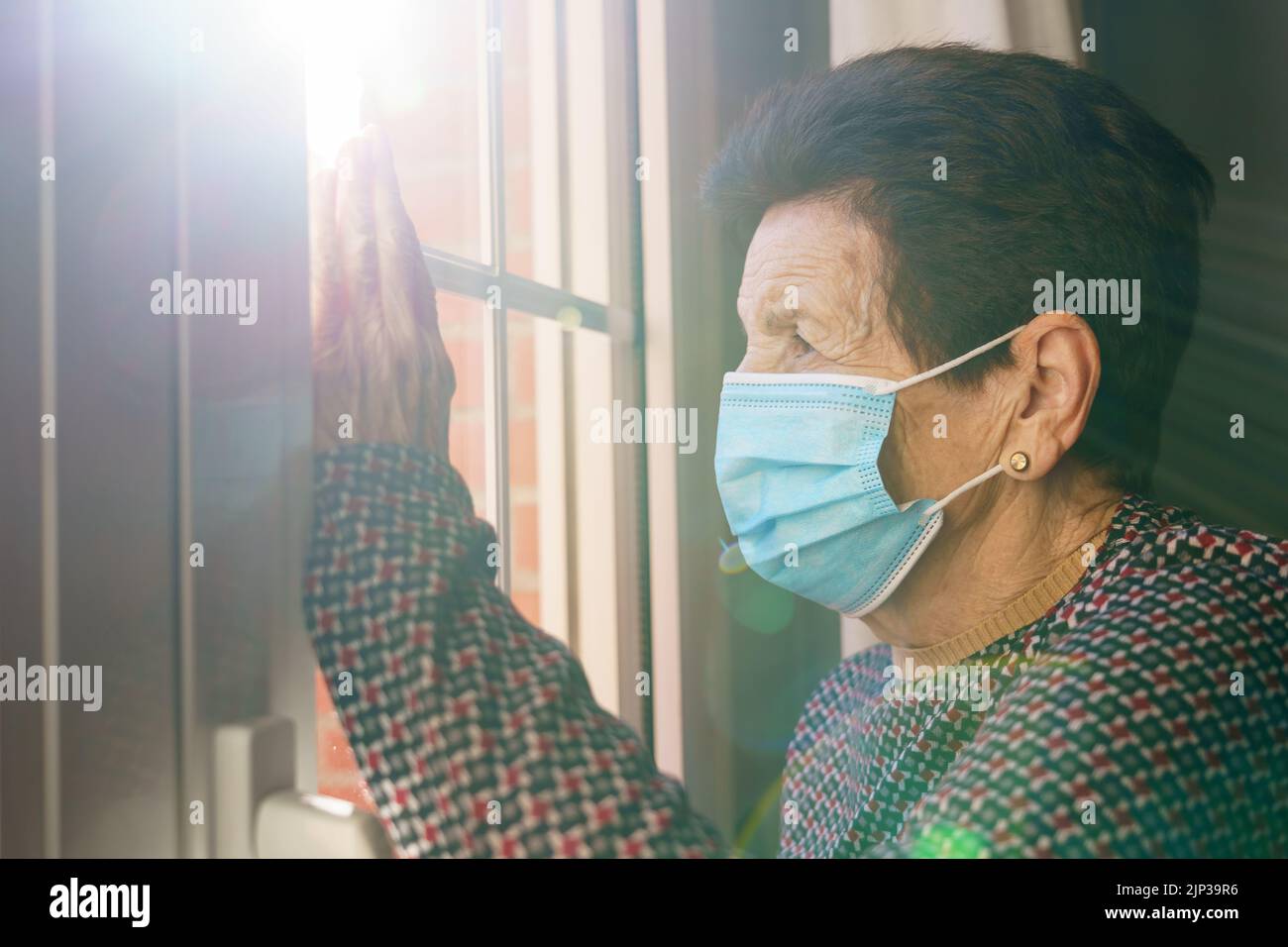 Elderly woman with a mask inside her house looking out the window with fear of the outside, due to the Covid-19 coronavirus. Stock Photo