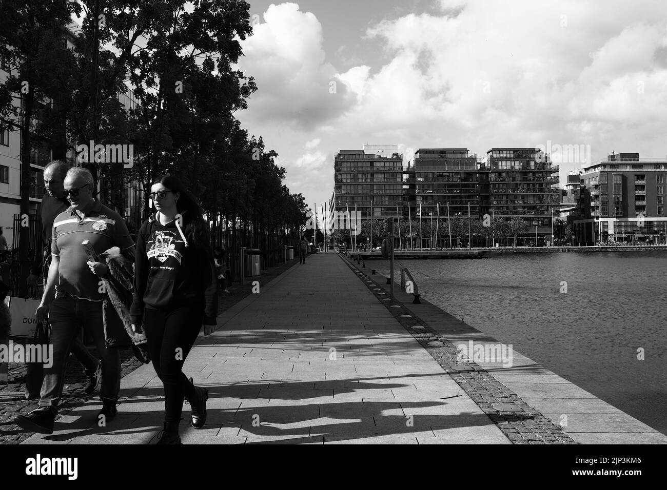 A grayscale of people walking by water at Grand Canal Dock with modern building in the background Stock Photo