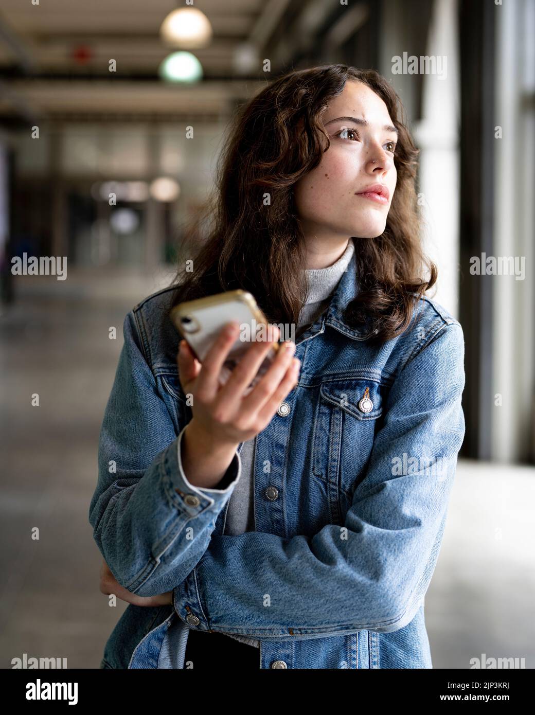 Unposed Portrait of Young Woman Using Smart Phone | Denim Jacket | Hallway Stock Photo