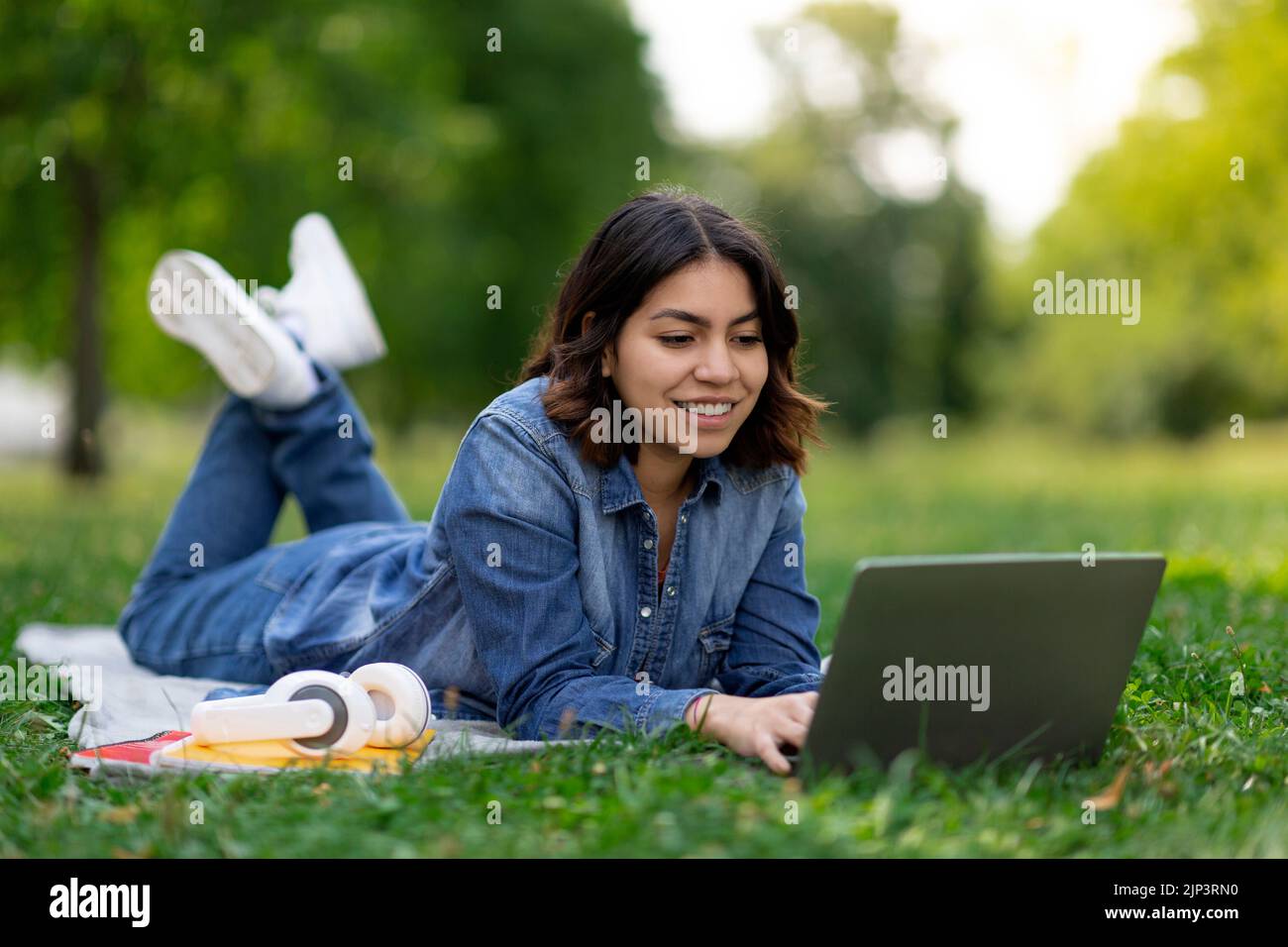 Smiling arab female student using laptop while lying on lawn outdoors Stock Photo