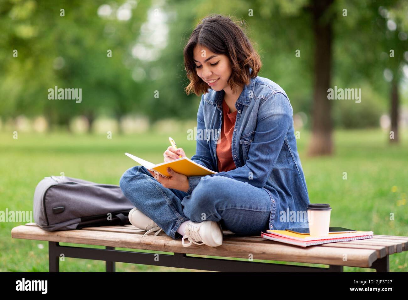Arab female student writing in notebook while sitting on bench in park Stock Photo