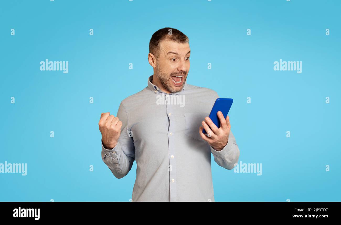 Emotional Man Holding Phone Shaking Fists Over Blue Background Stock Photo