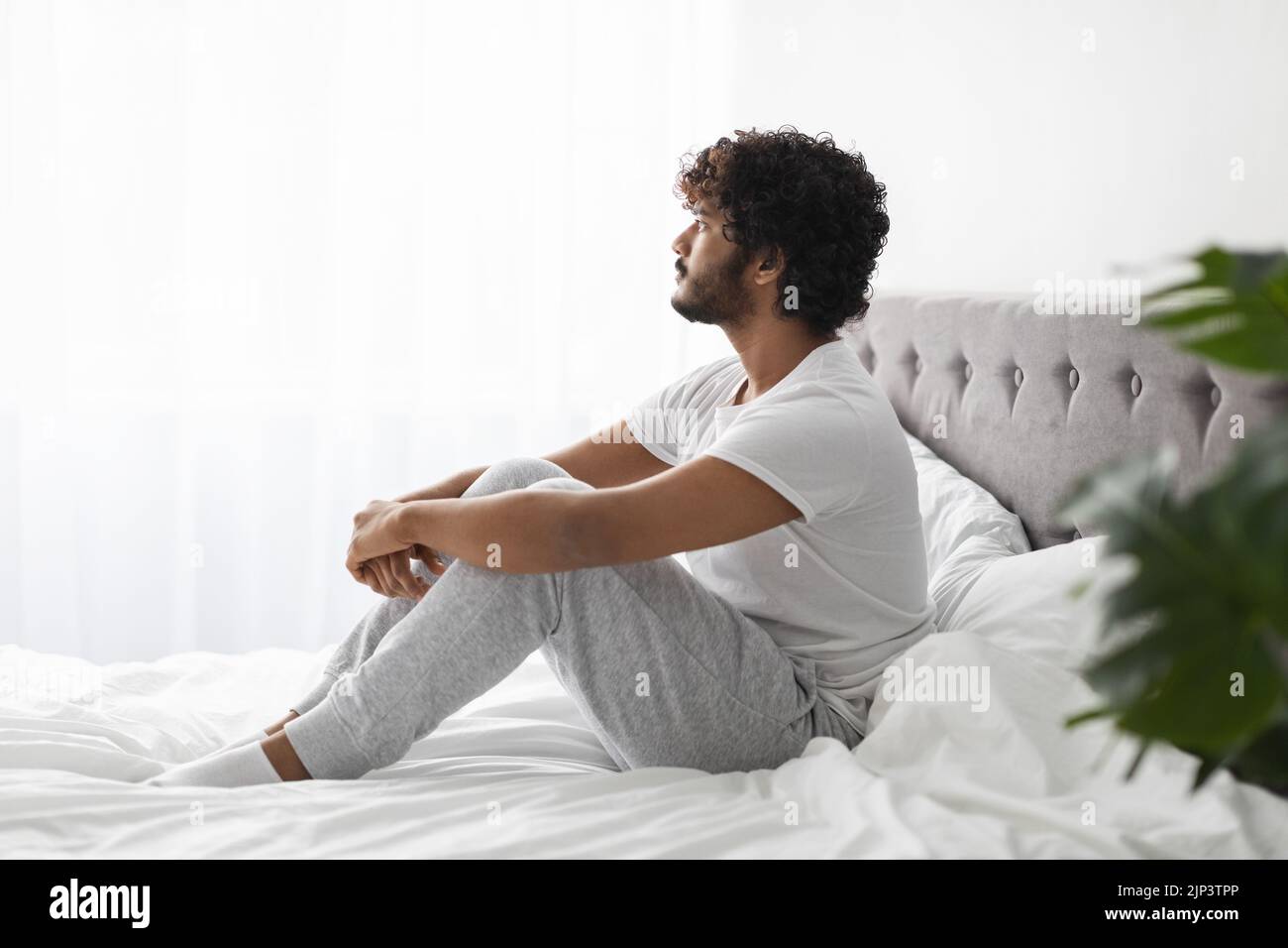 Pensive indian guy sitting on bed at home Stock Photo