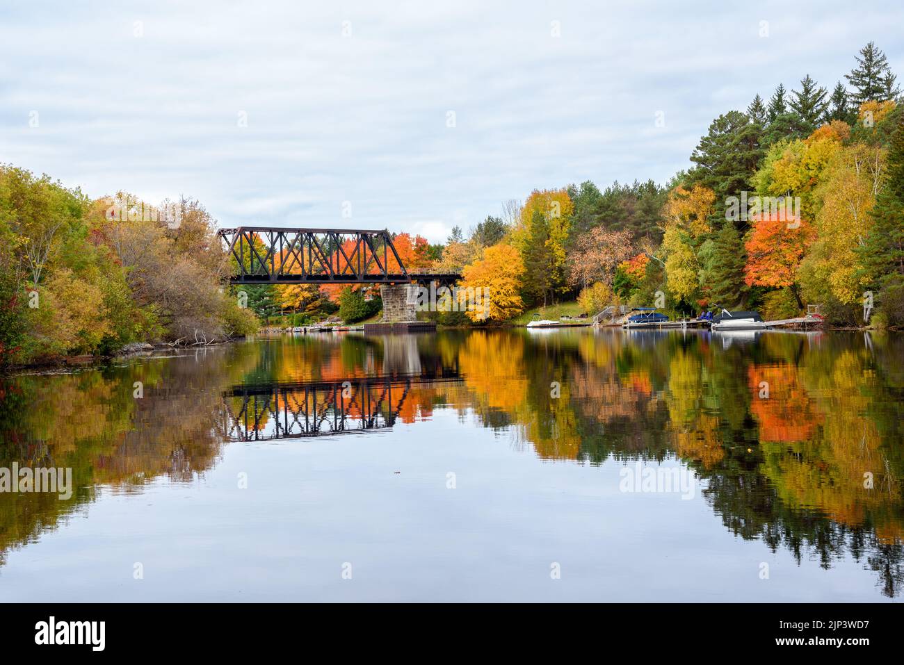 View of a steel railroad bridge spanning a river with forested banks at the peak of autumn colours. Reflection in water. Stock Photo