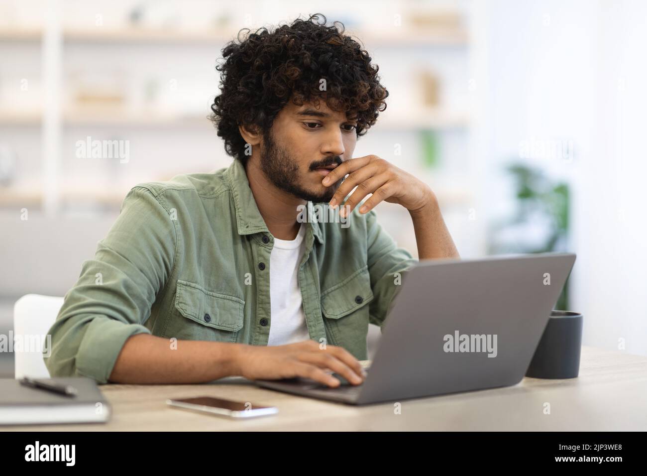 Pensive dark-skinned guy sitting at workdesk in front of laptop Stock Photo