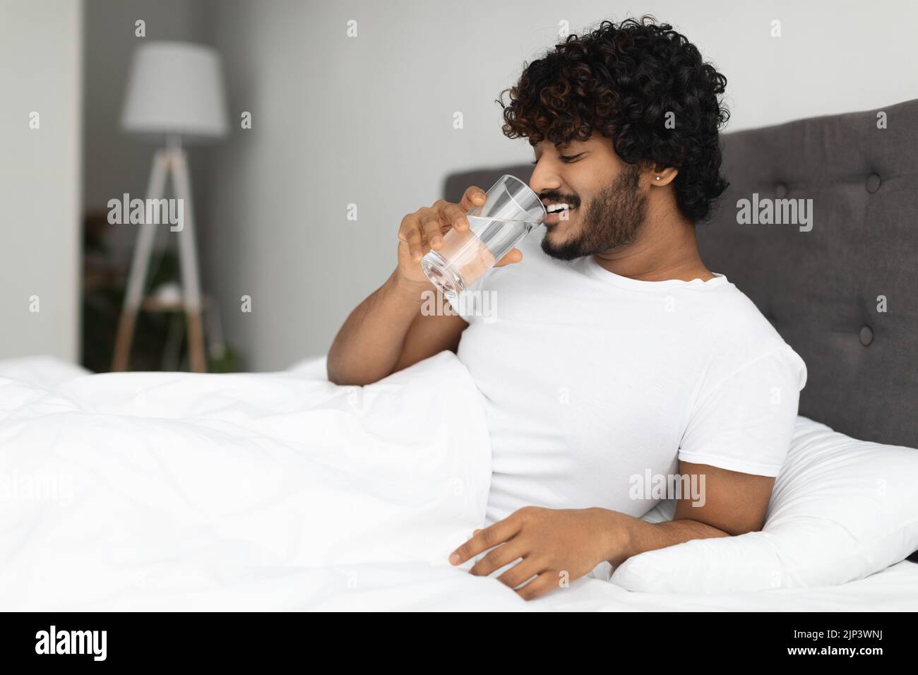 Happy young indian man holding glass of water in bed Stock Photo