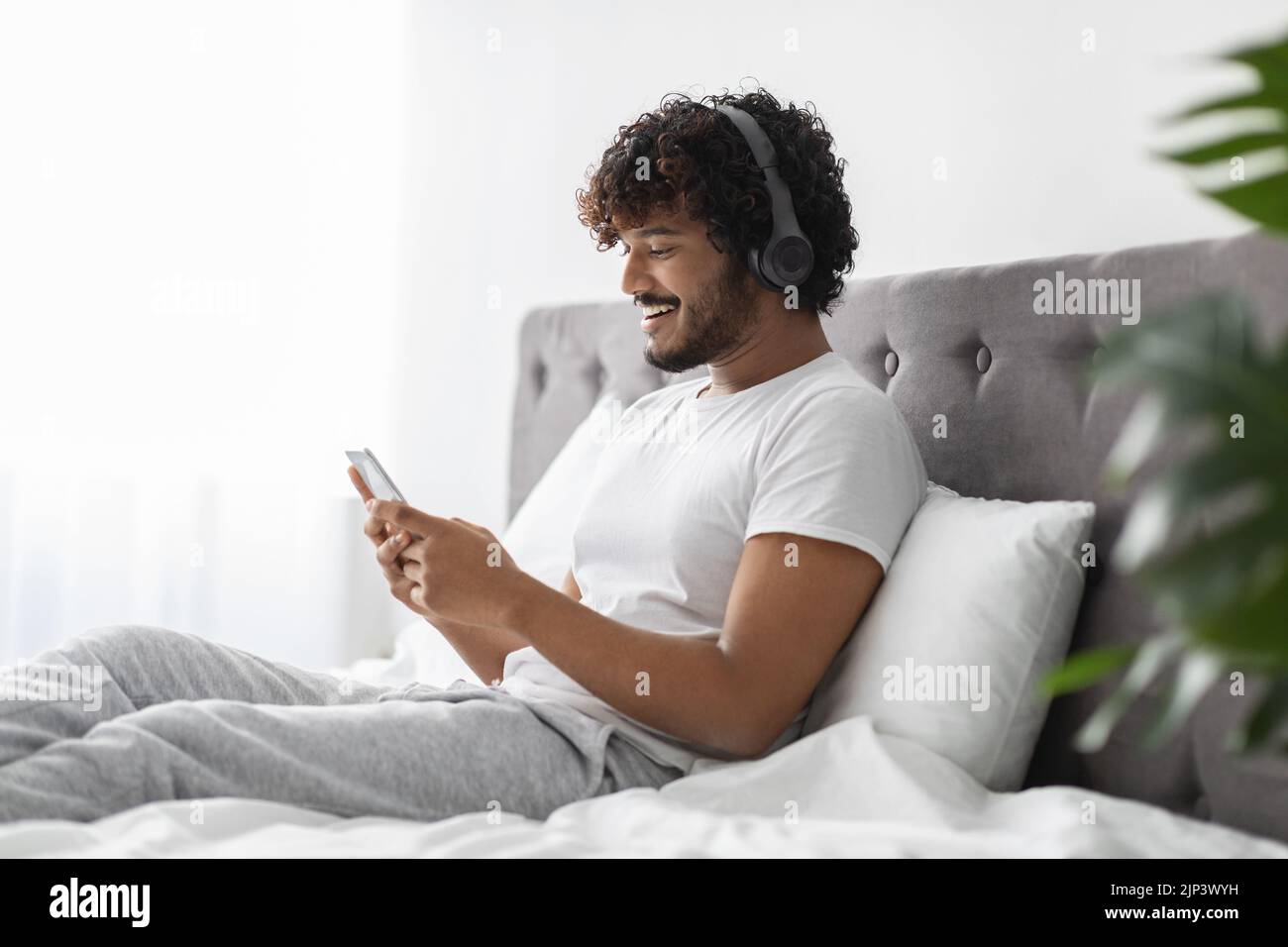 Cheerful hindu guy using cell phone and headphones in bed Stock Photo