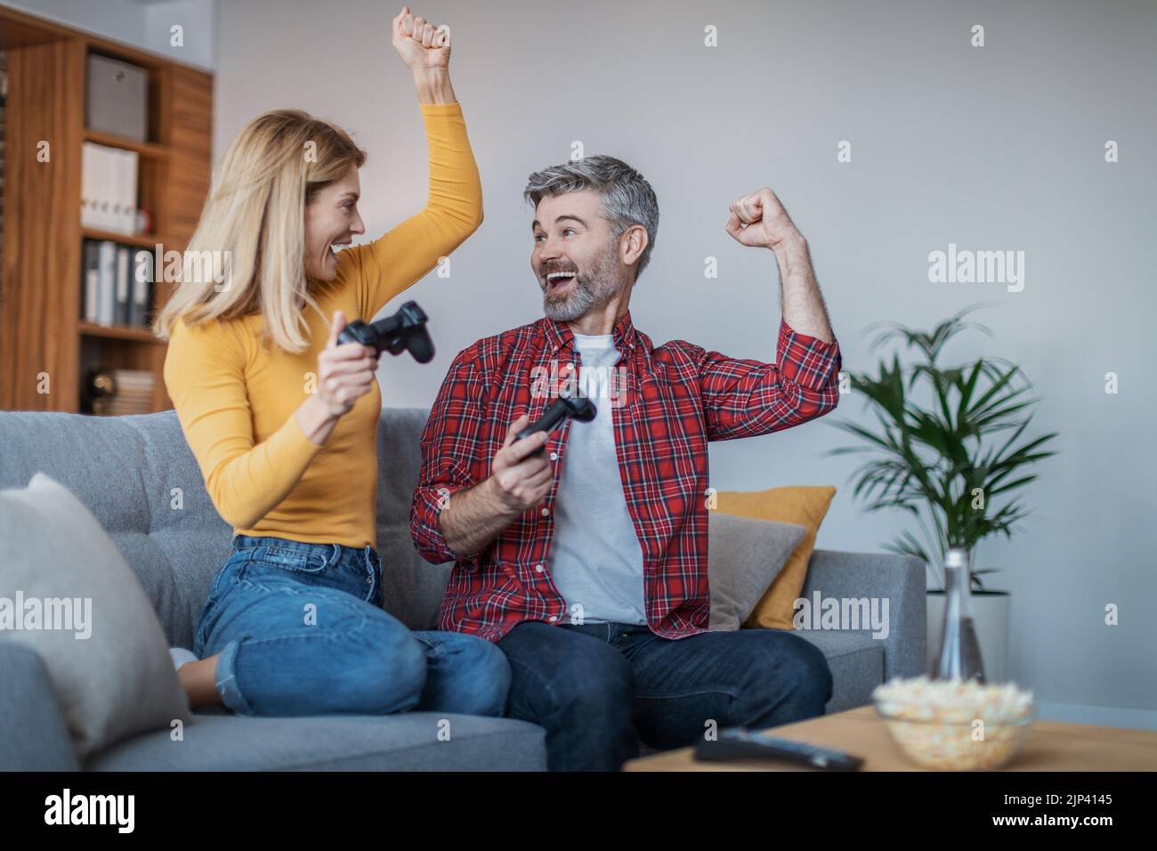 Happy excited adult european man and woman with joysticks enjoy victory, sit on sofa in living room Stock Photo