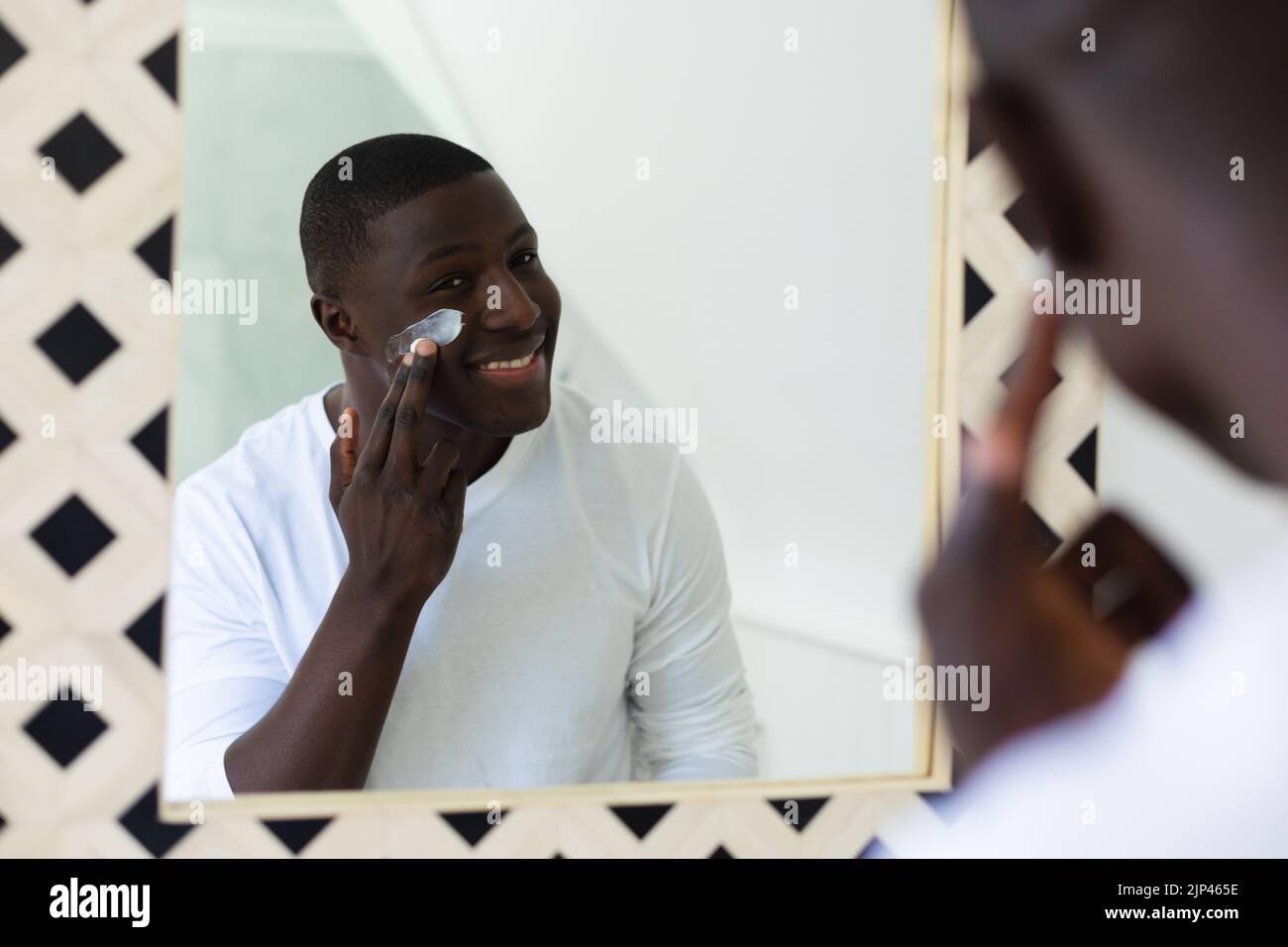 Smiling african american man applying face moisturiser looking in bathroom mirror Stock Photo