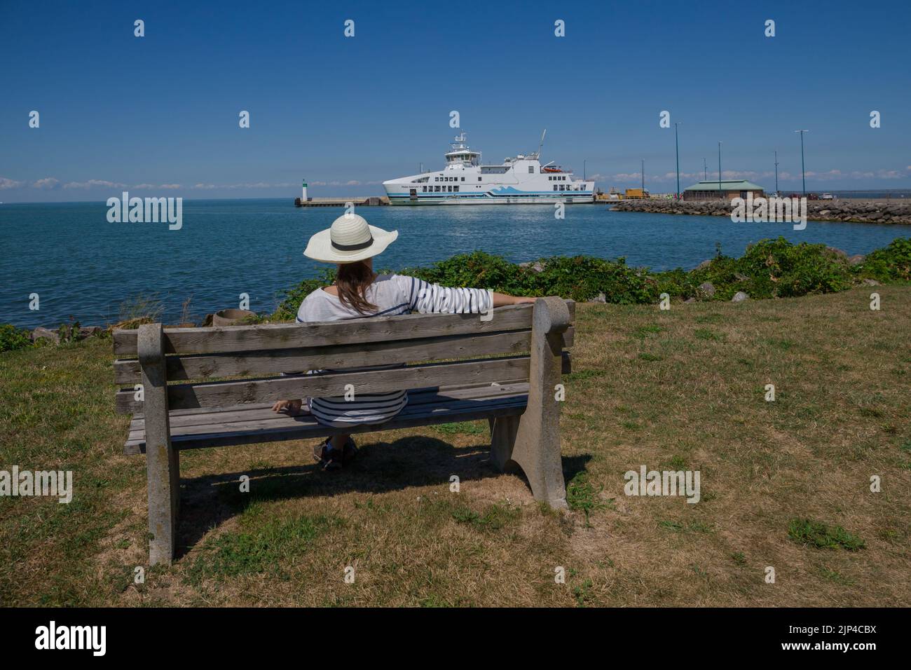 A woman in a hat seat on bench and looking of a ferry ship from the shore. Water transportation cruise liner Stock Photo