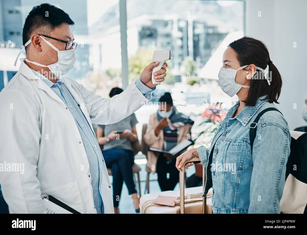 Covid traveling with a doctor taking temperature of a woman wearing a mask in an airport for safety in a pandemic. Healthcare professional with an Stock Photo