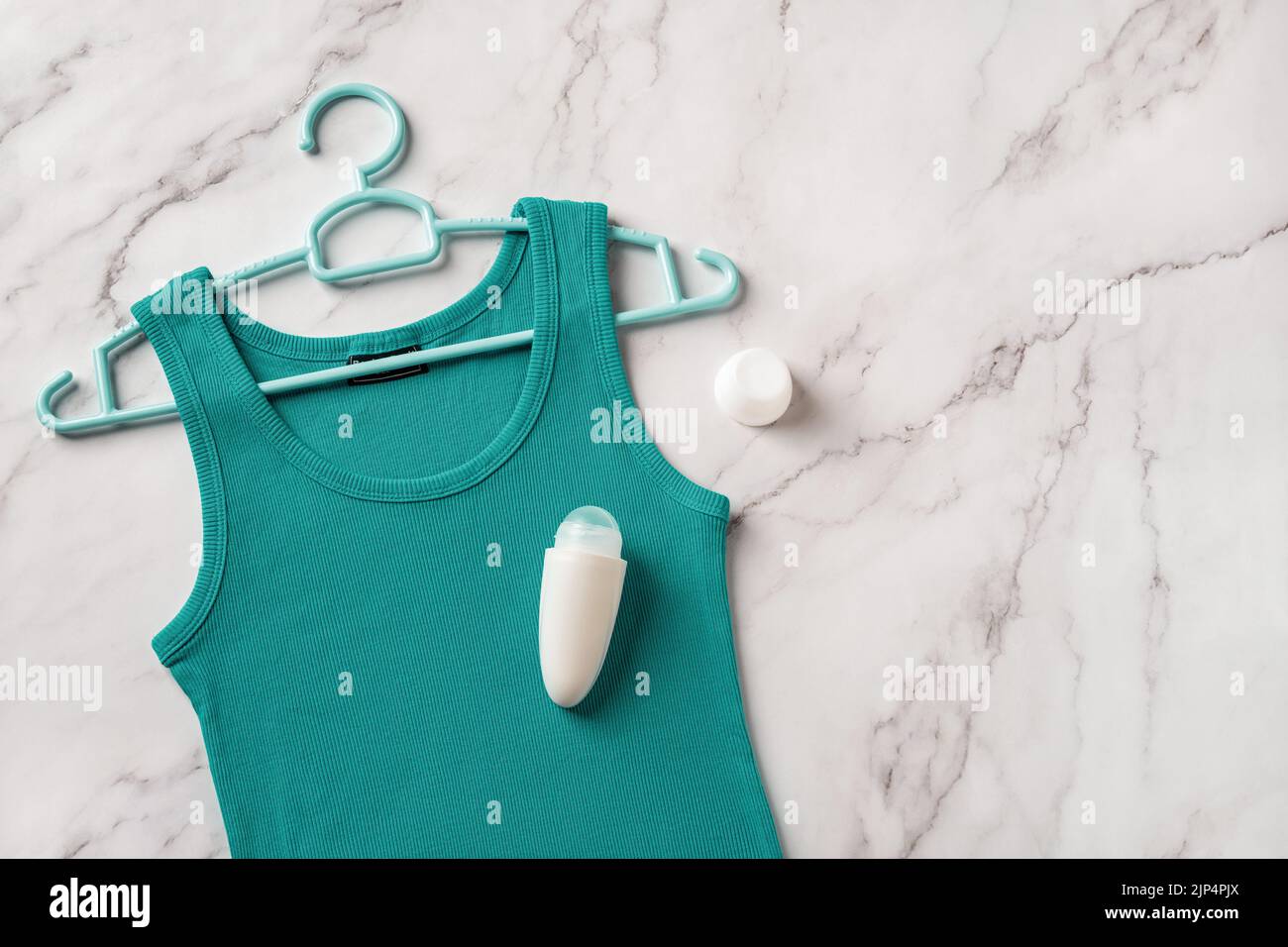 Roll on antiperspirant on a teal tank top over marble background. Linen jersey tank on a hanger and roll-on deodorant to prevent body odour. Hygiene. Stock Photo