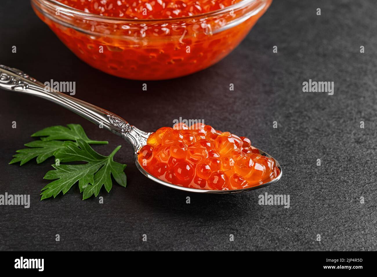 Salmon red caviar in a spoon on a black slate background. Vintage spoon full of trout caviar and parsley leaf close-up. Seafood fish delicacy. Stock Photo