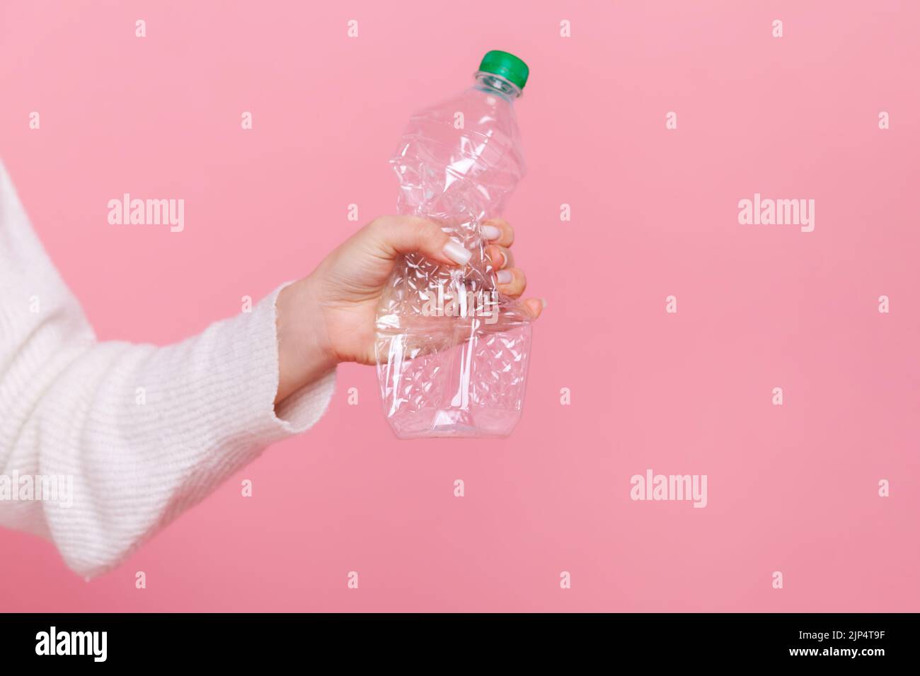 Female hand holding plastic bottle with green cap, sorting her rubbish, worrying about environment, wearing white casual style sweater. Indoor studio shot isolated on pink background. Stock Photo