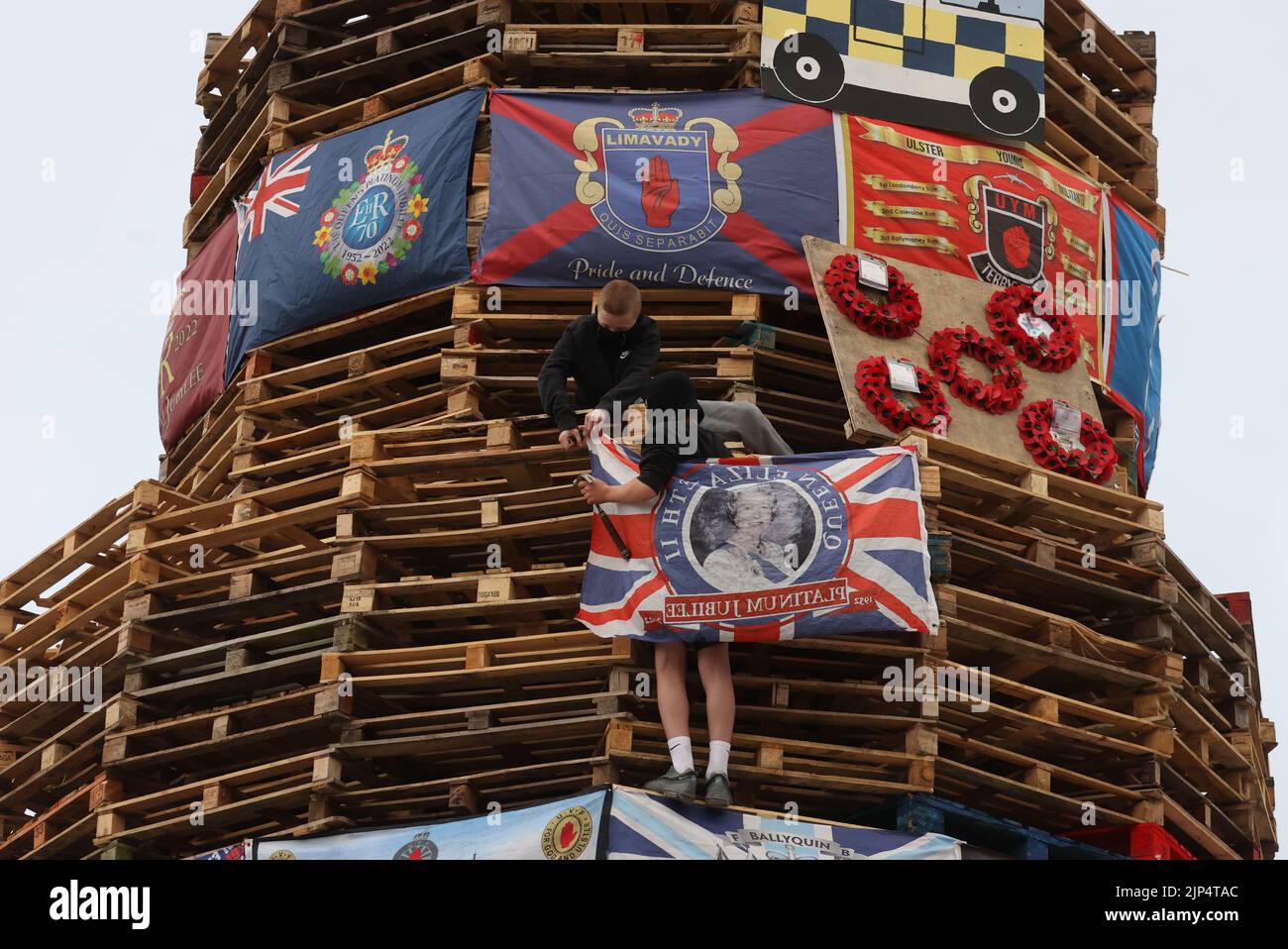 Young boys fix a union flag bearing the Queen's face to a bonfire prior to it being lit to mark the Catholic Feast of the Assumption in the Bogside area of Londonderry, Northern Ireland. Picture date: Monday August 15, 2022. Stock Photo